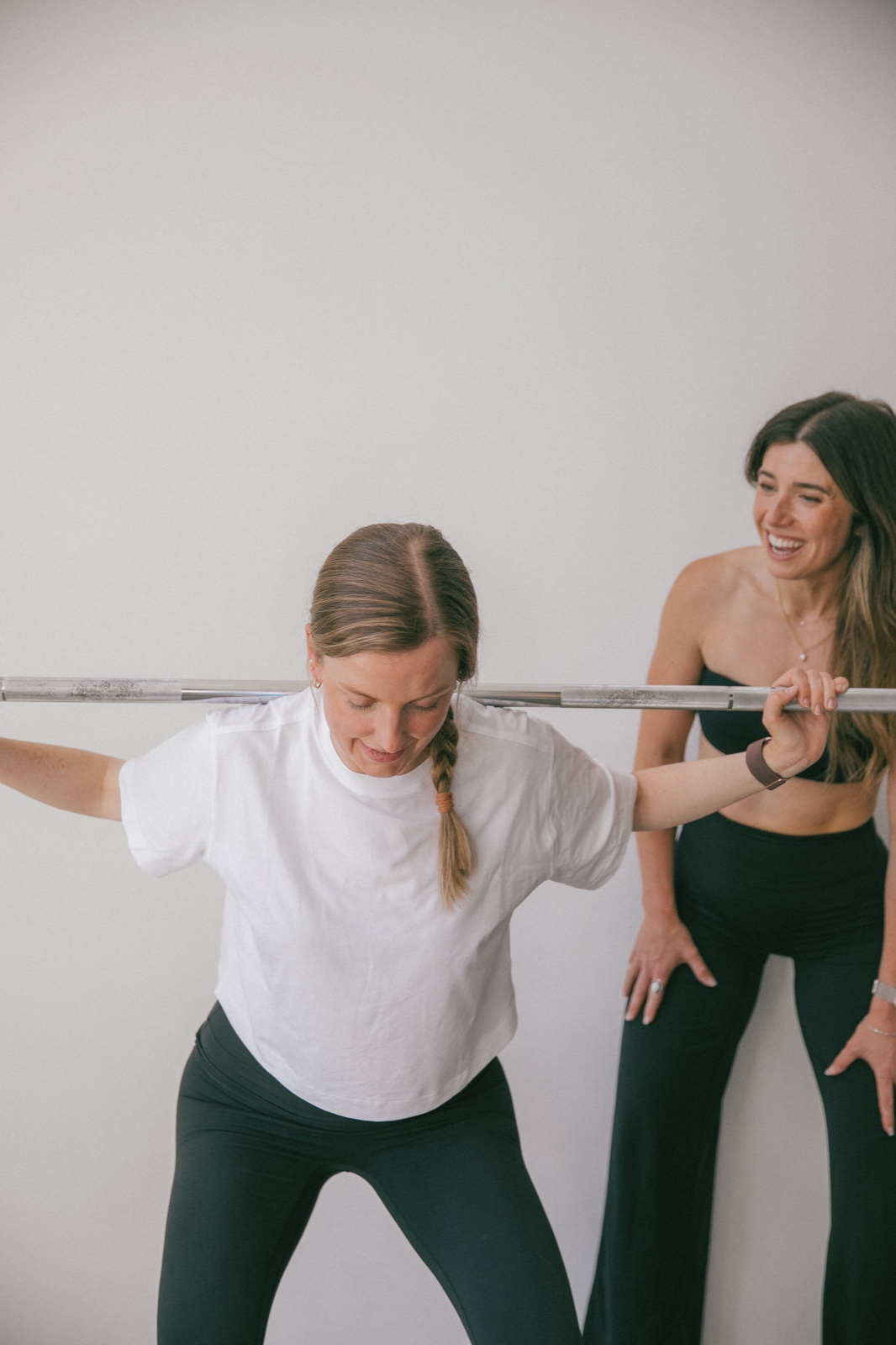 A client steadying a barbell across her shoulders while Alex coaches beside her, laughing