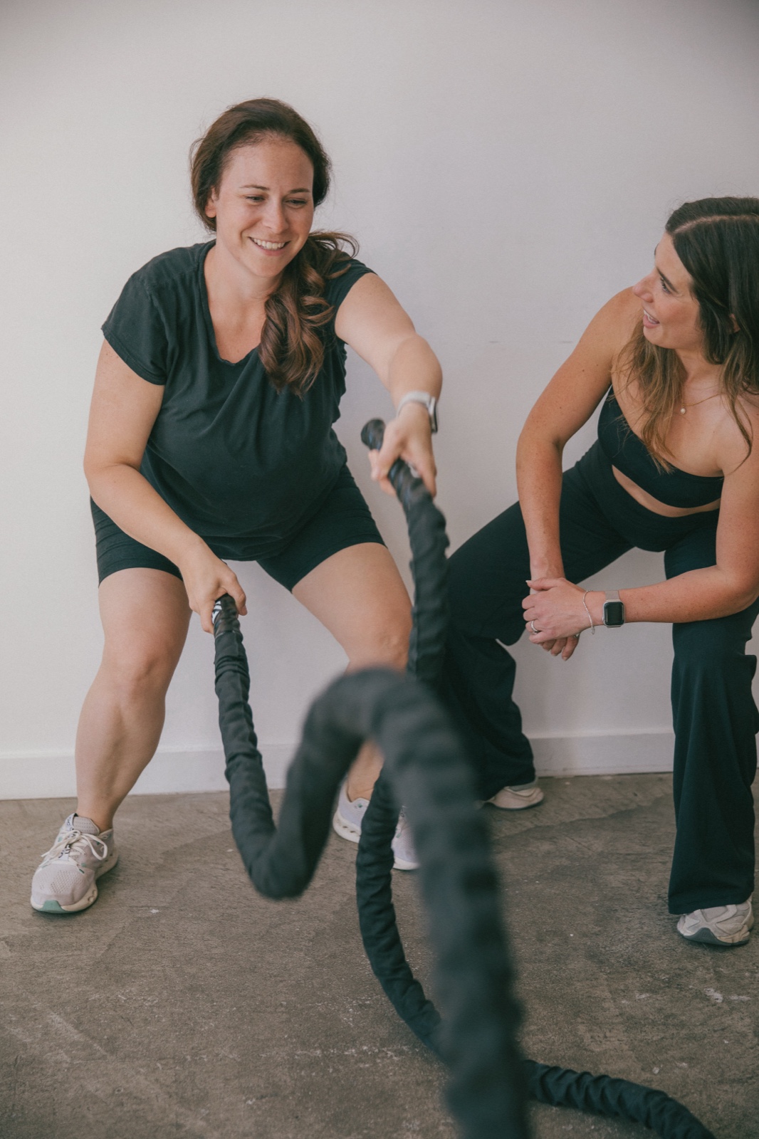 A client working battle ropes, laughing, while Alex crouches beside her watching the work